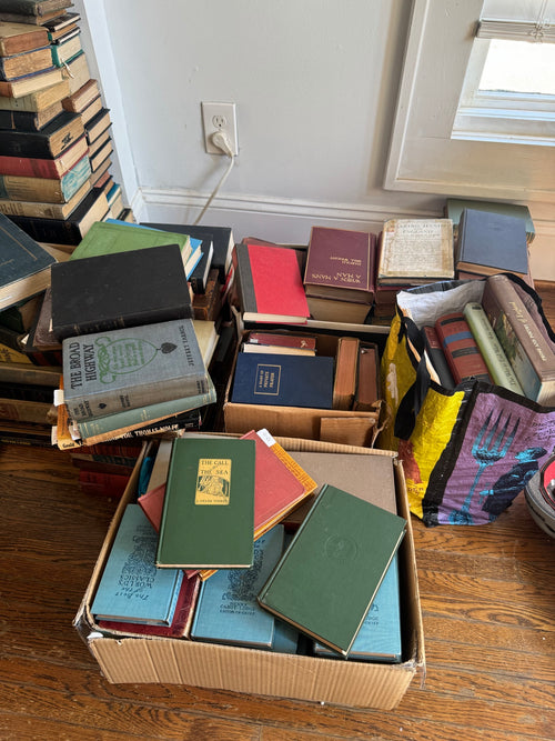 Cardboard boxes and wooden shelf displaying large antiquarian book collection sorted by color and size, white wall with electrical outlet visible