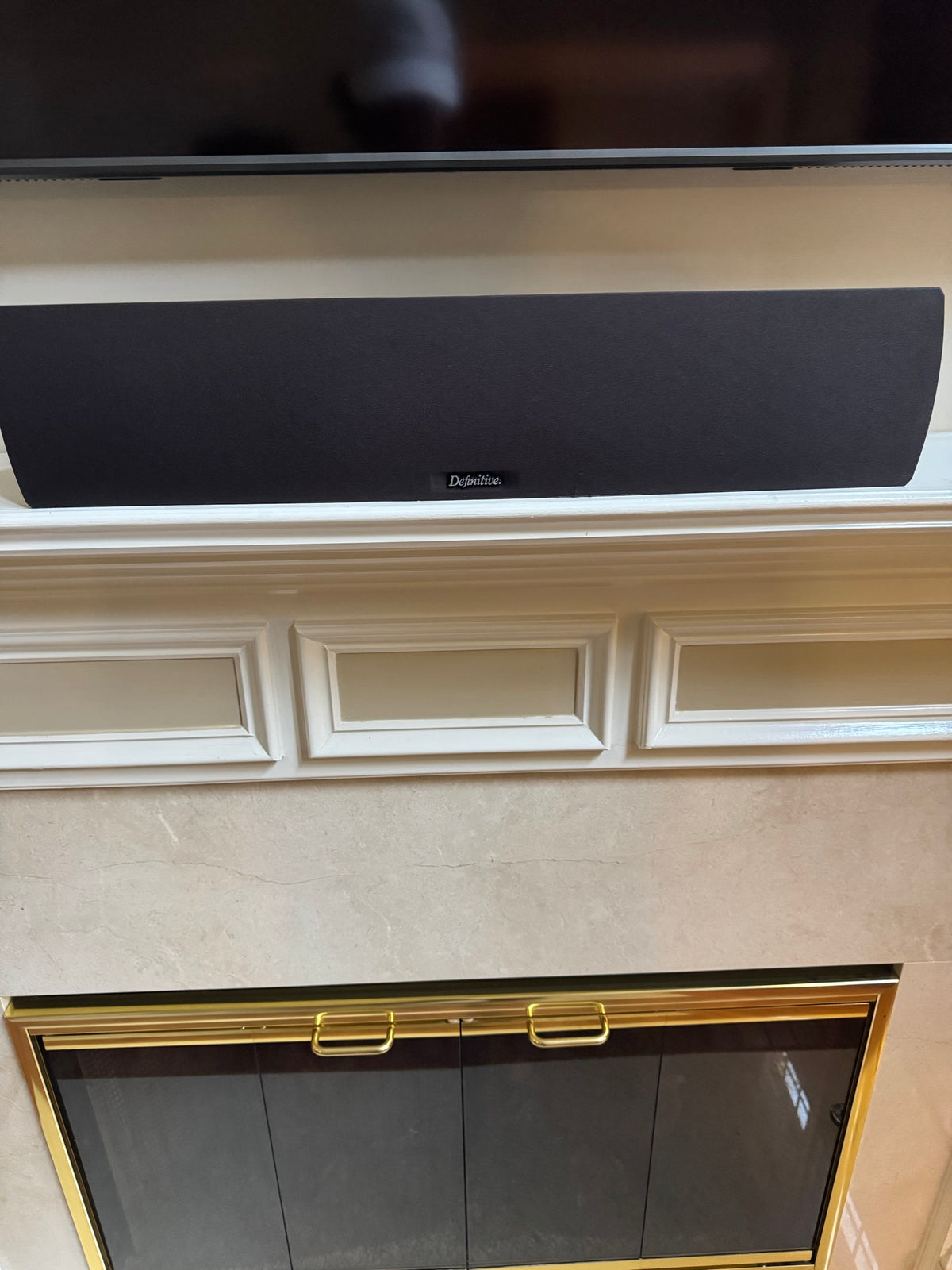Definitive Technology matte black center channel speaker mounted on a mantel below a wall-mounted TV, with a fireplace and brass-trimmed glass doors below
