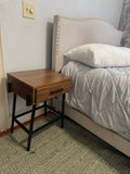 Wood one-drawer nightstand on a black metal base next to a bed