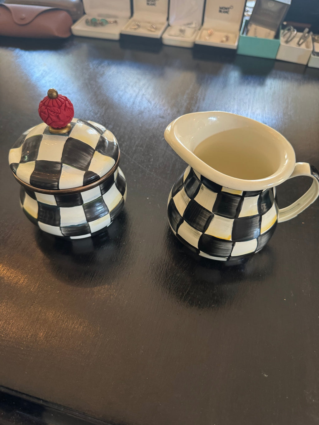 Black and white checkered sugar bowl with lid topped by red finial, and matching cream pitcher displayed on dark wood surface