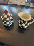 Black and white checkered sugar bowl with lid topped by red finial, and matching cream pitcher displayed on dark wood surface