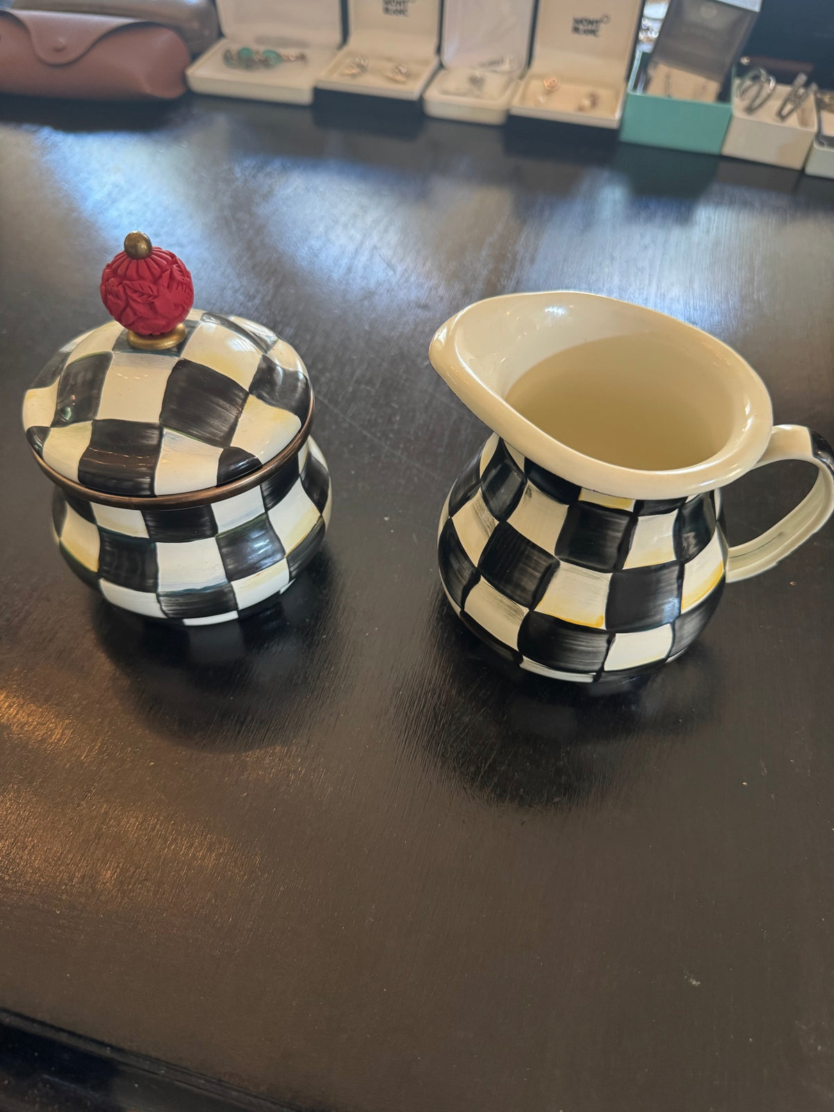 Black and white checkered sugar bowl with lid topped by red finial, and matching cream pitcher displayed on dark wood surface