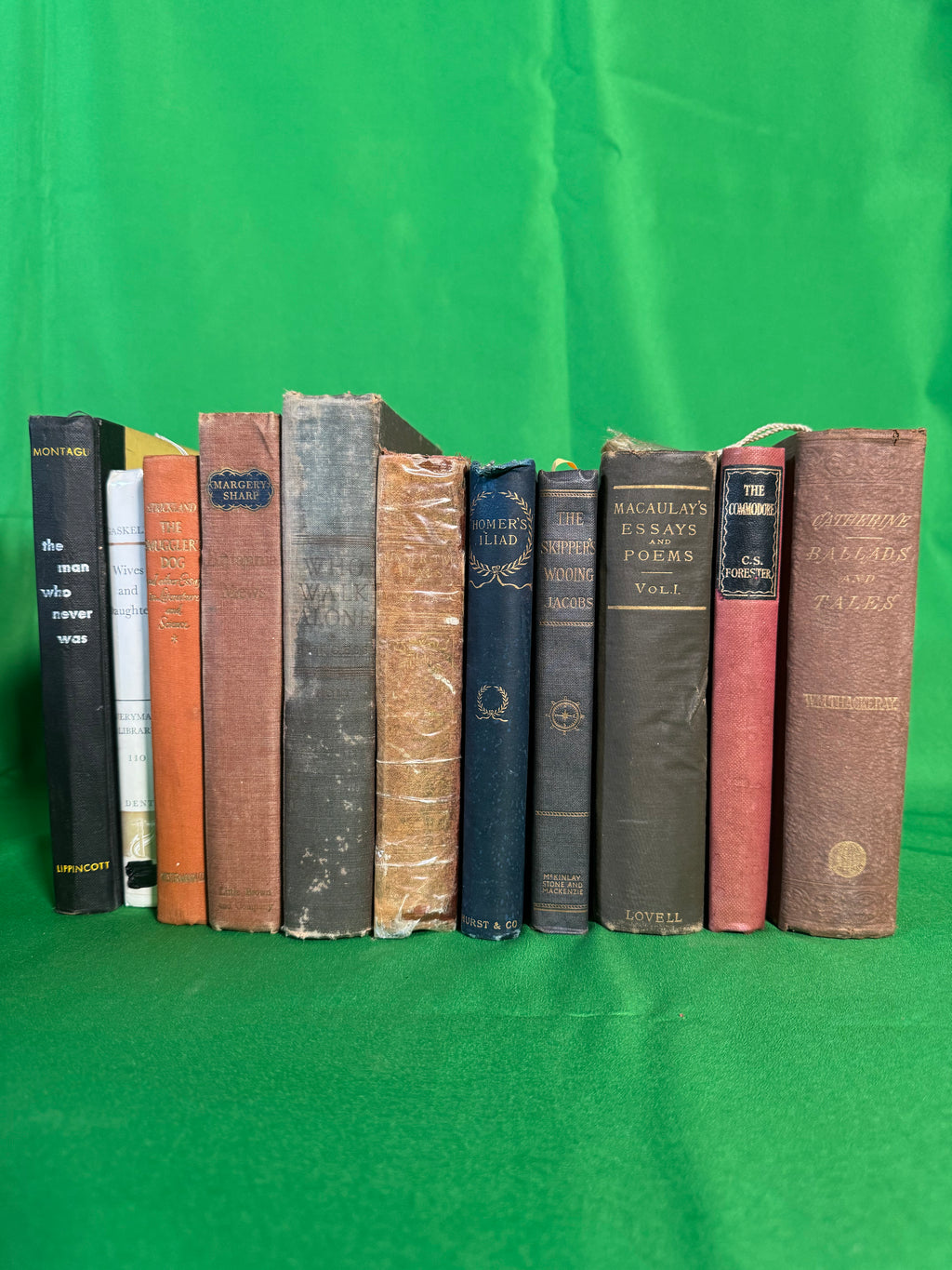 Vintage hardcovers stood spine-out on green cloth, including The Commodore by C. S. Forester, Macaulay's Essays and Poems, Thackeray's Catherine and Ballads and Tales, and The Man Who Never Was