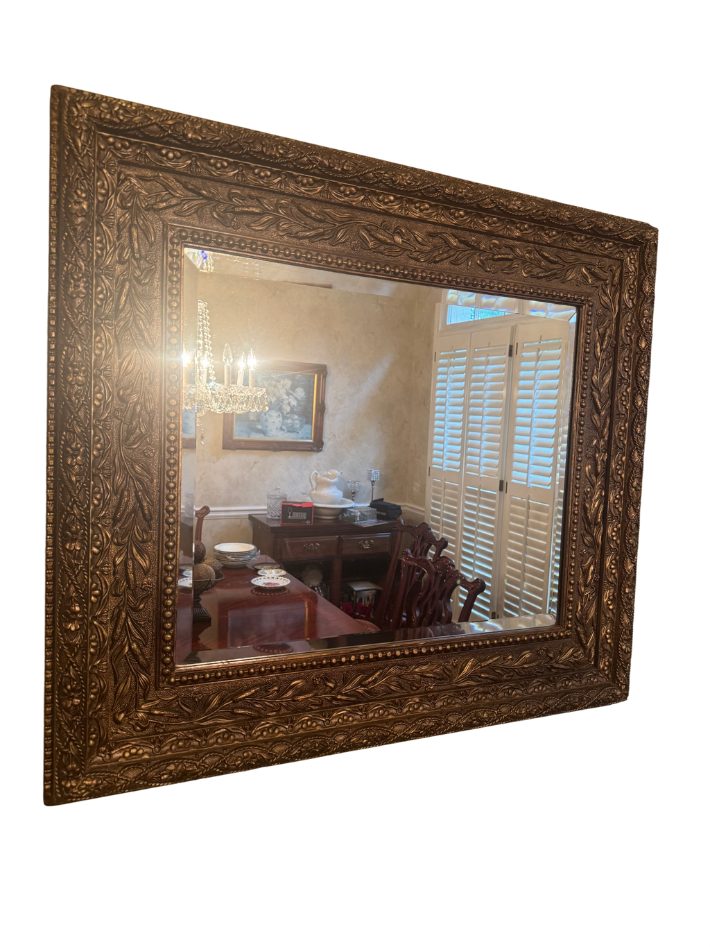 Rectangular wall mirror with ornate embossed gilt frame and bevelled glass, hanging on a textured wall, reflecting a dining room