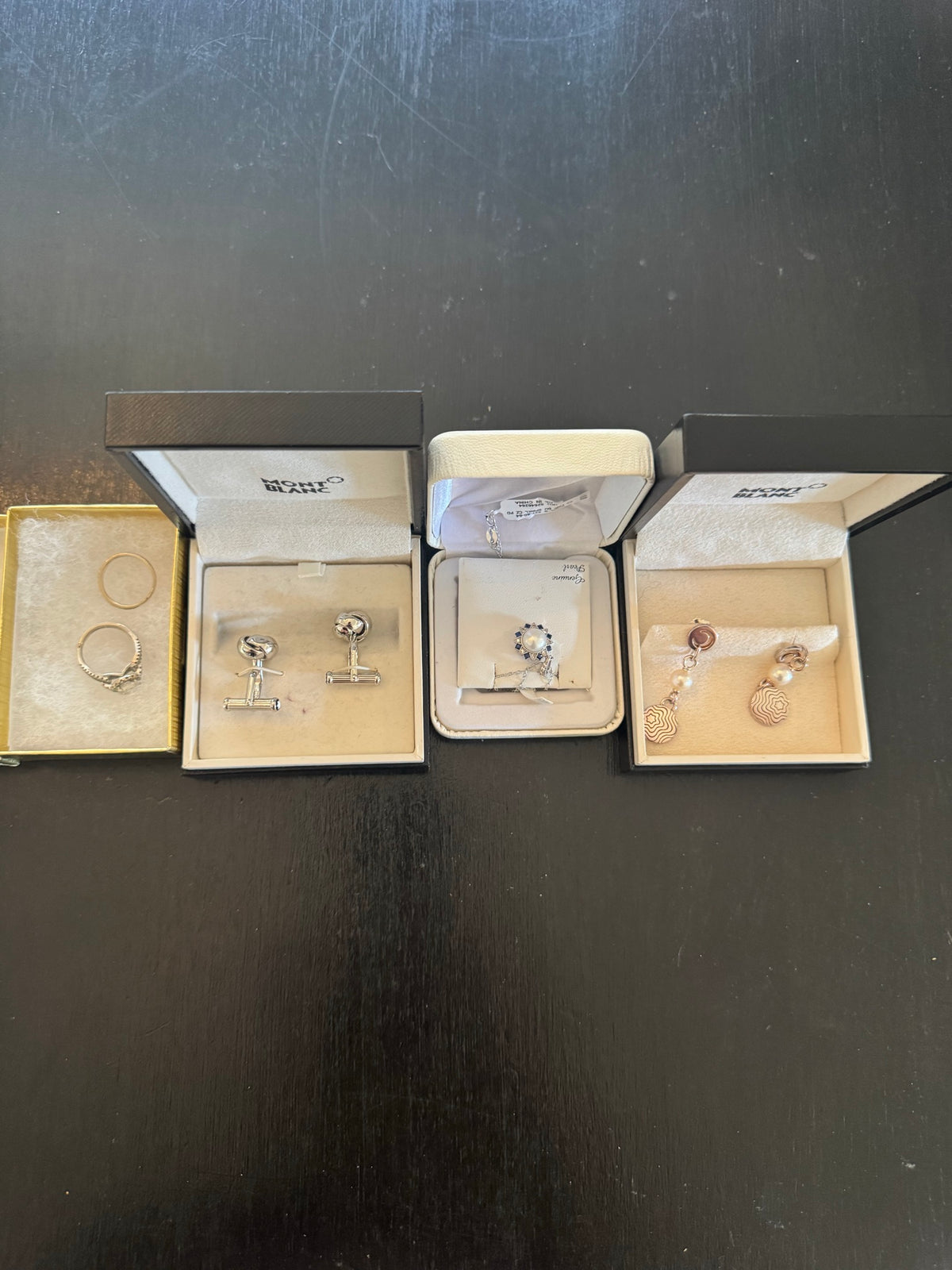 Flat lay of four Montblanc branded boxes and one gold box containing various jewelry pieces on dark surface