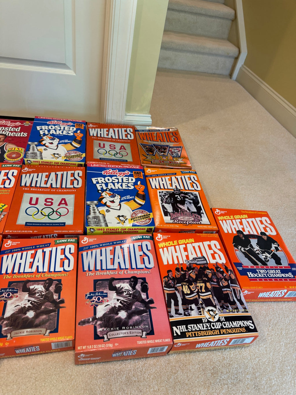 Multiple unopened cereal boxes displayed on carpeted stairs, showing Wheaties, Frosted Flakes with championship and Olympic Games commemorative designs