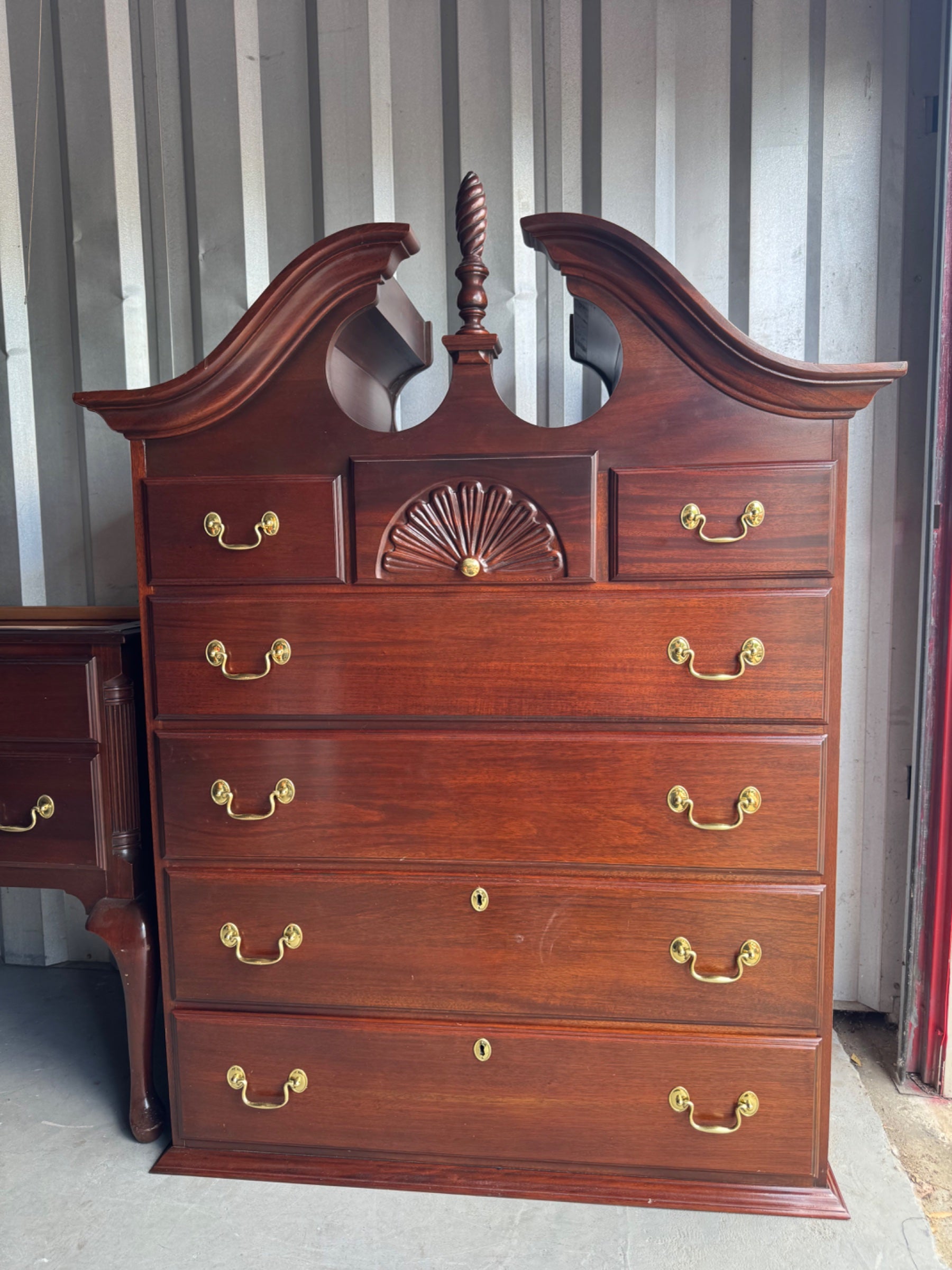Mahogany highboy chest viewed from front, showing ornate carved pediment with scrolled wings and central finial