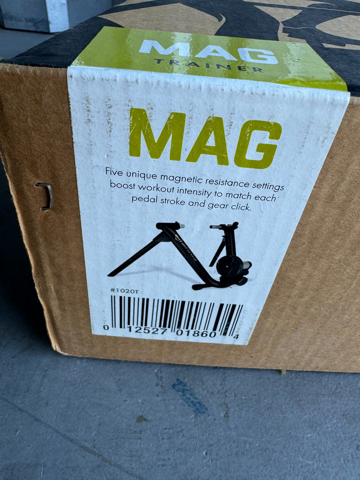 Cardboard box side showing MAG Trainer product label with green banner, magnetic resistance bike trainer silhouette, barcode visible