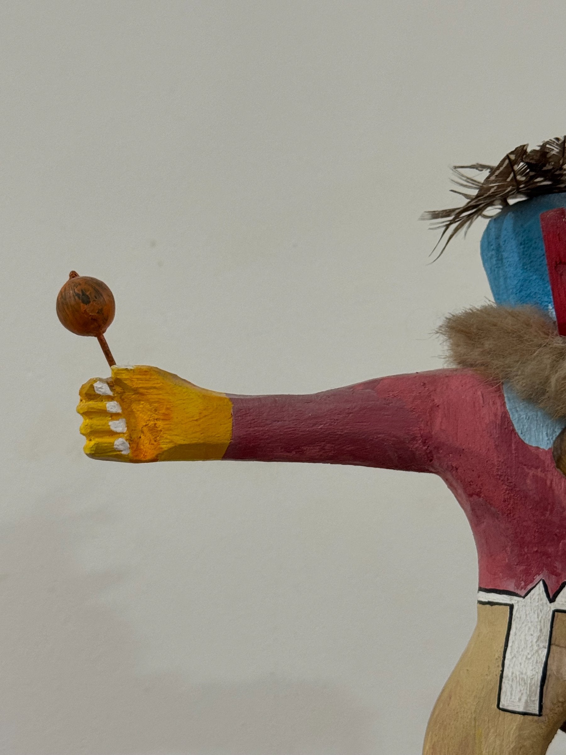 Detail of right arm extended outward with yellow painted hand holding round brown ball on stick