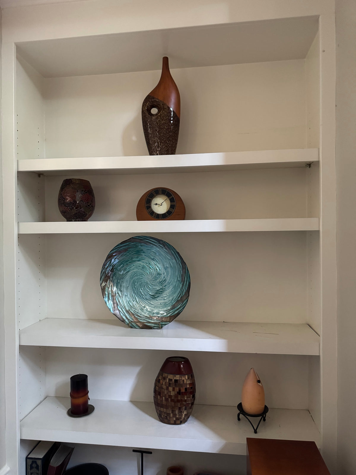 Bookshelf display with wood and ceramic fish-form vase, mosaic vase, round wood-cased clock, teal swirl art glass plate, mosaic tile vase, pillar candle, and teardrop vase on stand