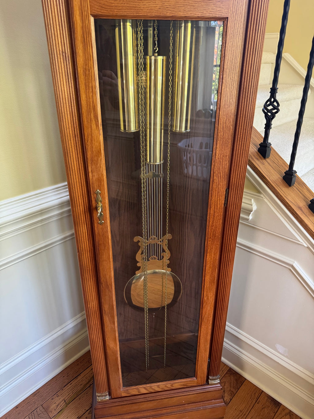 Interior view through glass door showing brass-cased weights, lyre pendulum, and chain mechanism against dark background panel
