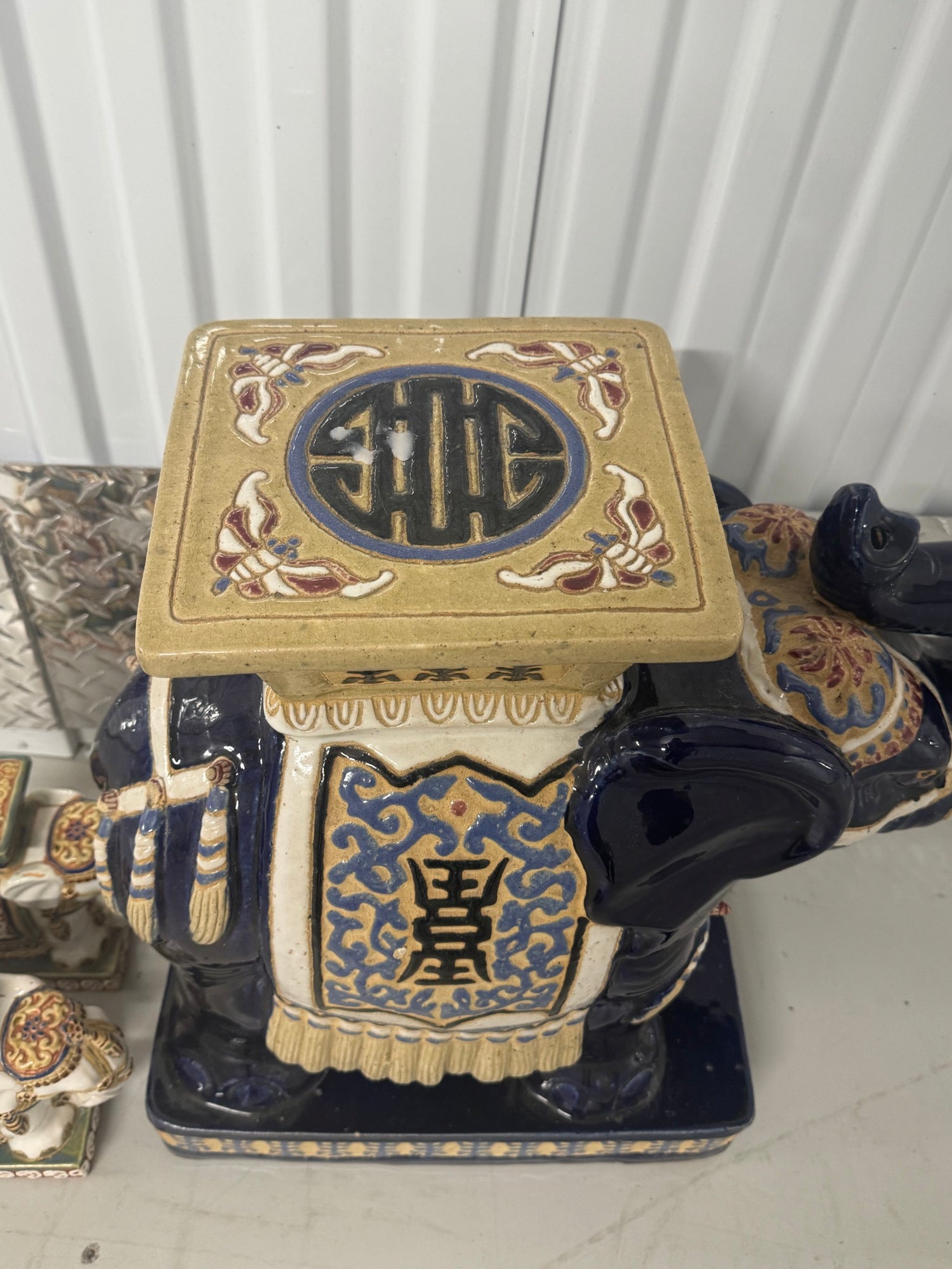Close-up of gold ceramic elephant stool top with blue oval medallion containing gilt characters, cream body with gold patterned saddle