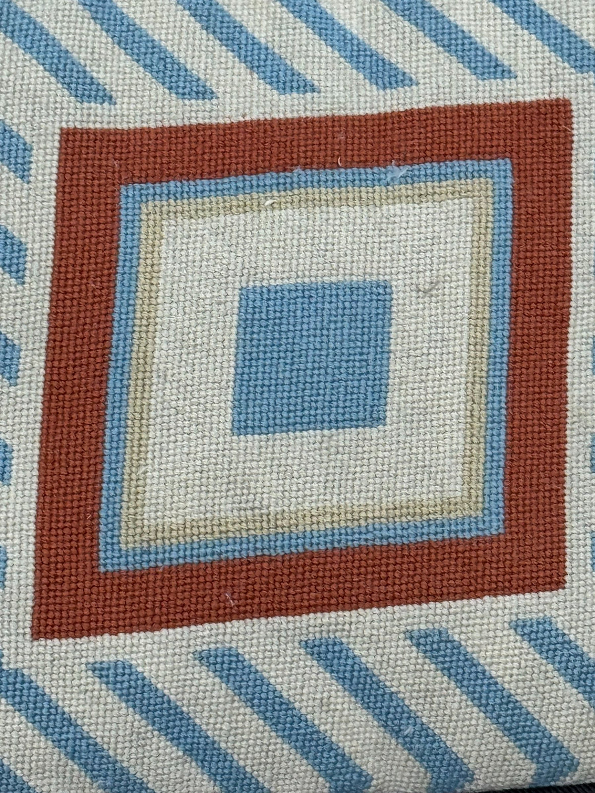 Close-up detail of needlepoint front showing concentric squares in rust, cream, light blue and darker blue with diagonal striped background pattern