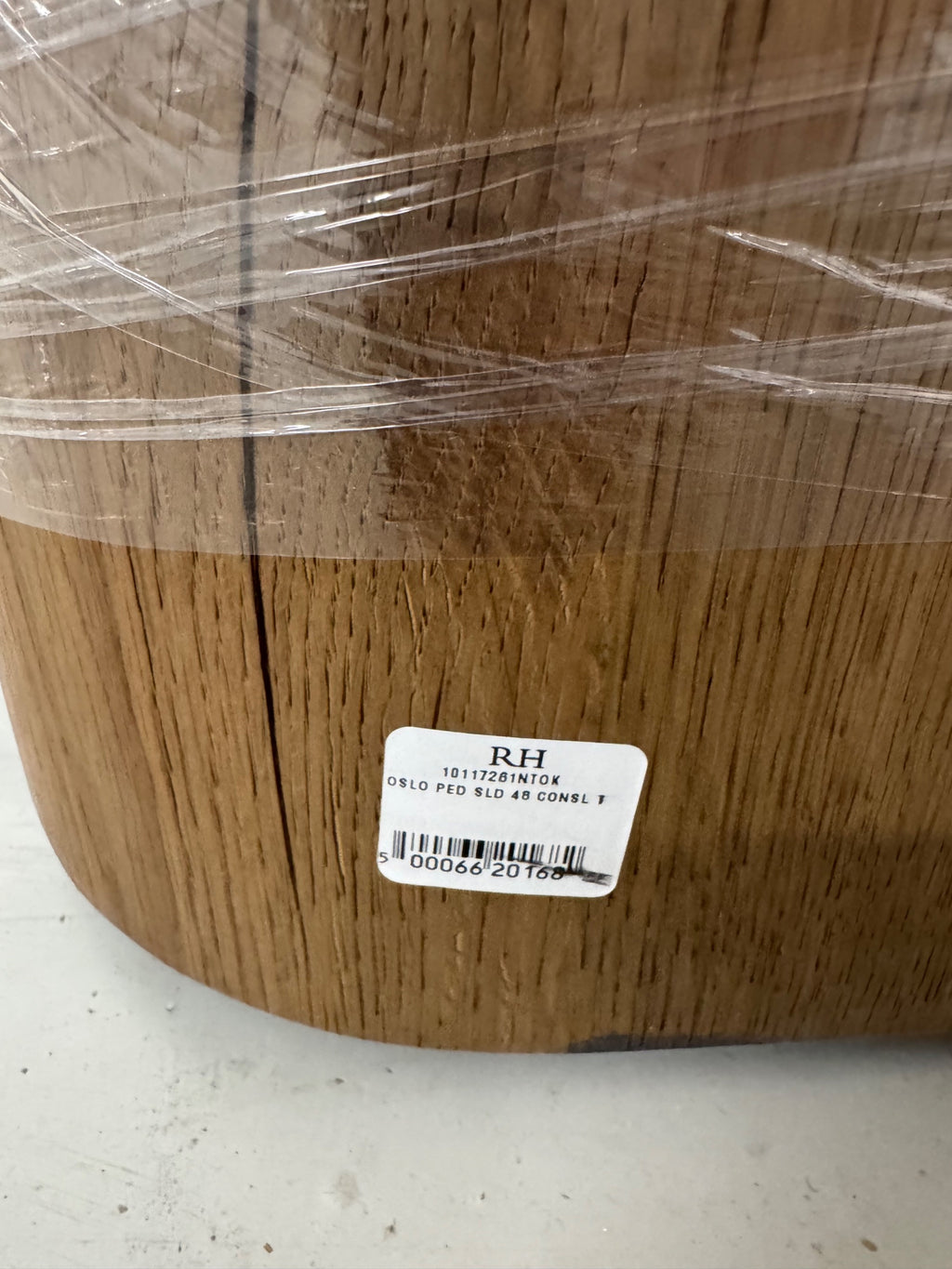 Close-up of walnut wood grain and vertical slats on pedestal base, white label reading 'RH 10117261NTOK OSLO PED SLD 48 CONSL T' with barcode