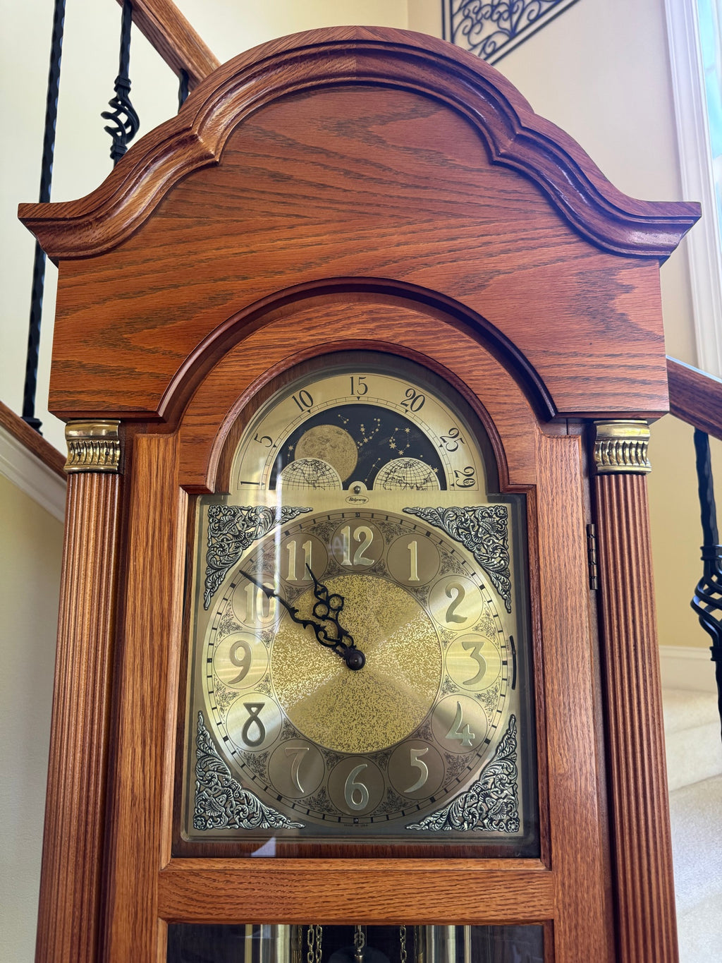 Close-up of clock face showing brass dial with moon phase complication above main twelve-hour dial and ornate corner spandrels