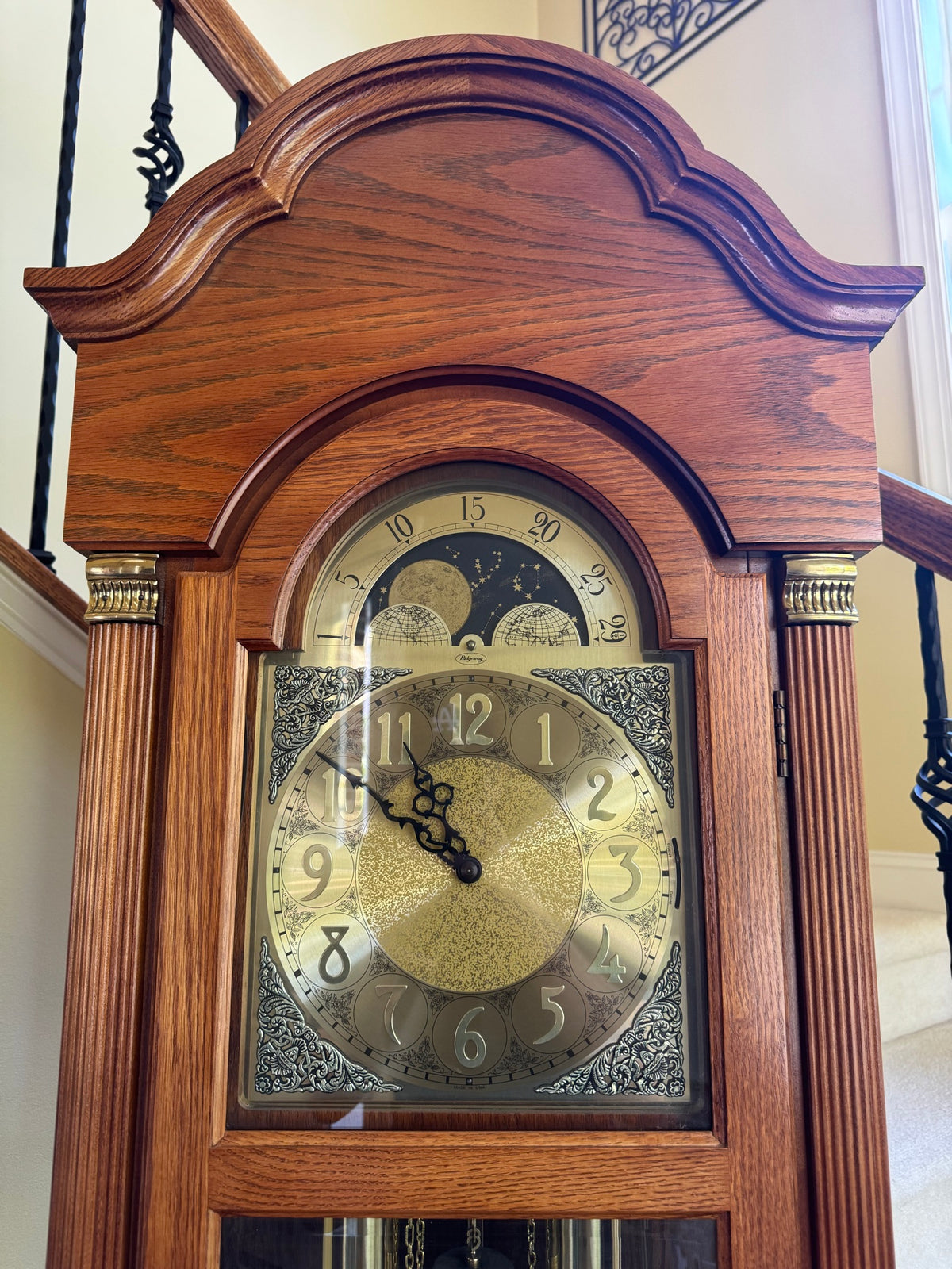 Close-up of clock face showing brass dial with moon phase complication above main twelve-hour dial and ornate corner spandrels