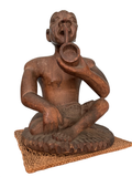 Seated carved wooden figure with tribal features, holding circular horn to mouth, woven mat base, front view