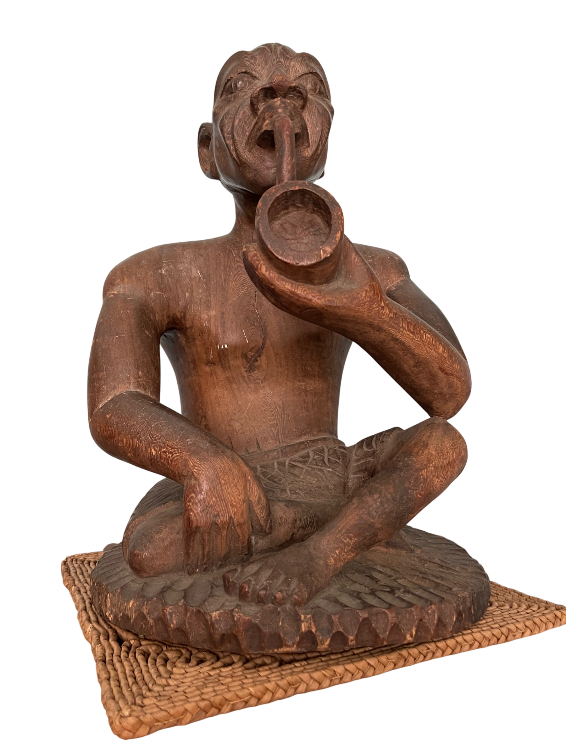 Seated carved wooden figure with tribal features, holding circular horn to mouth, woven mat base, front view
