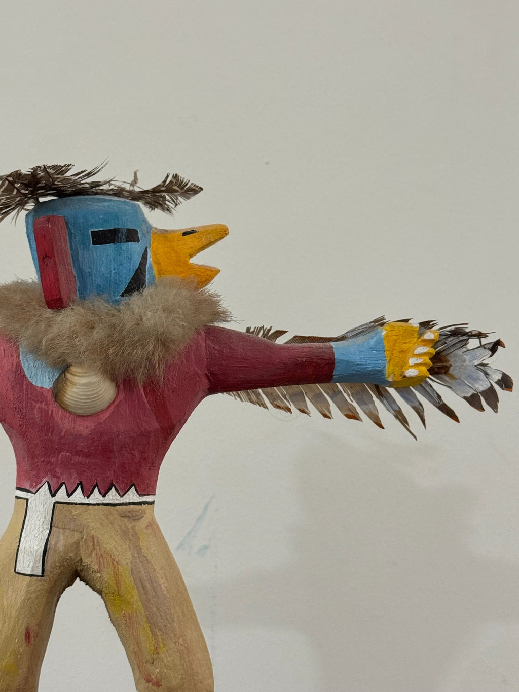 Side profile of kachina head showing blue and red painted sections, yellow beak, straw hair, and feathered wing detail