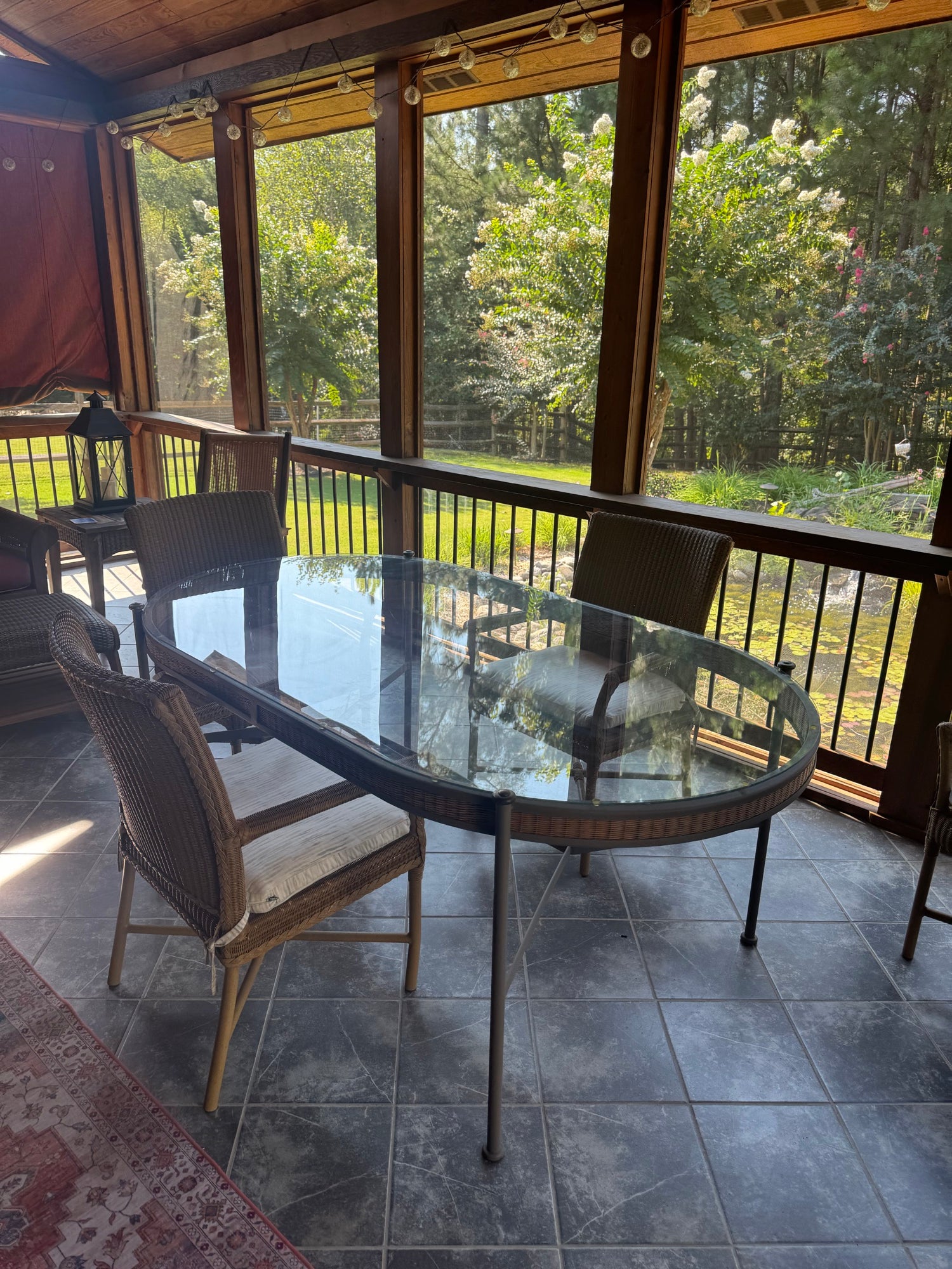 Oval glass-top dining table with dark metal frame and five woven resin wicker armchairs in sunlit screened porch