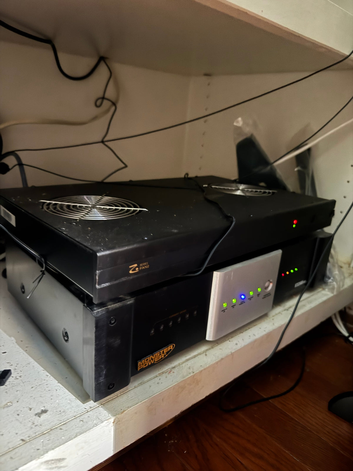 Stacked black rack electronics on a shelf, including a Monster Power branded power conditioner unit, a black cooling fan unit labeled Z Series Fan2, and a small silver panel with lit LED indicators.