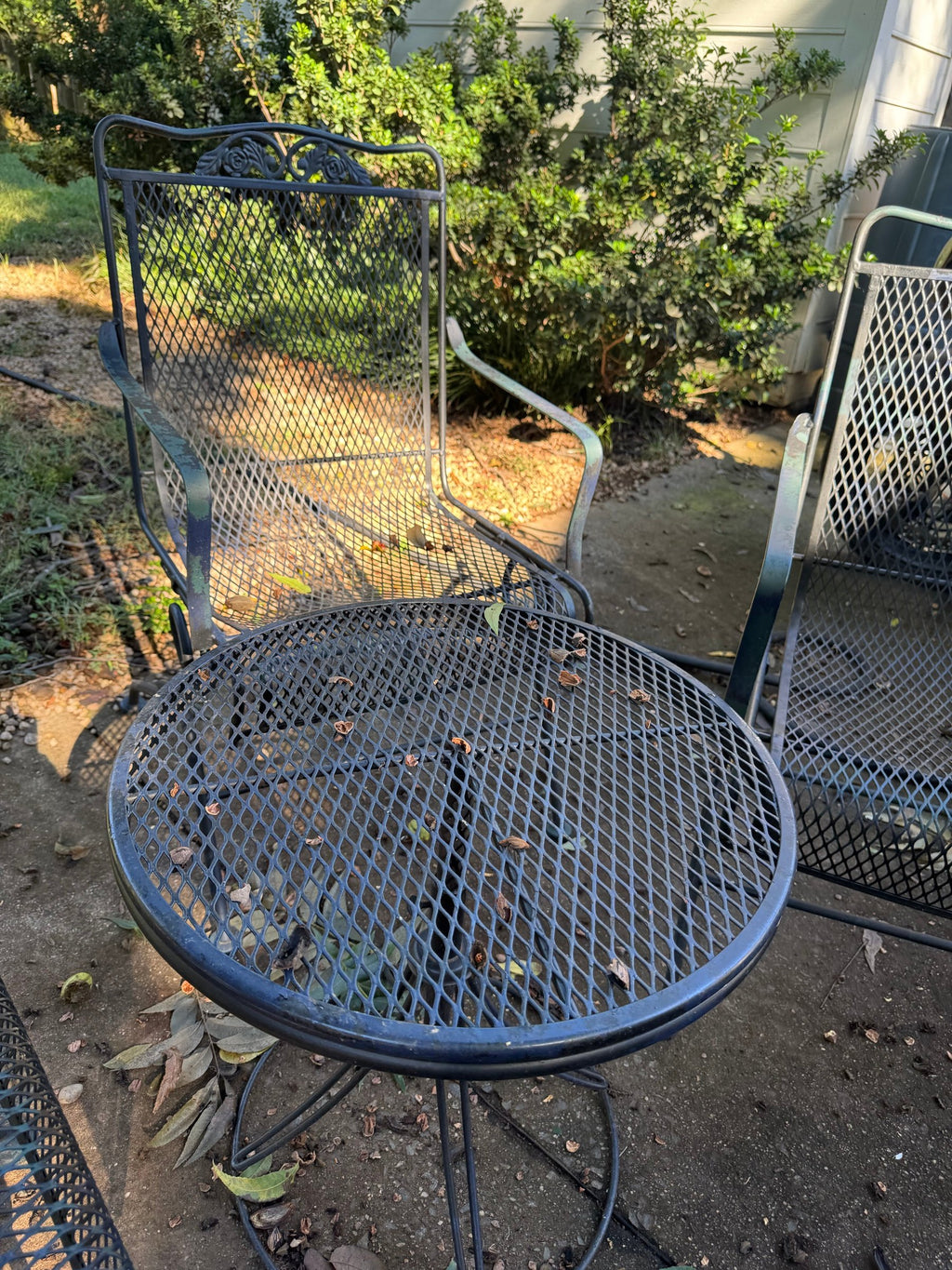 Two metal mesh spring chairs beside a round mesh-top table, one chair showing a lighter patina finish.