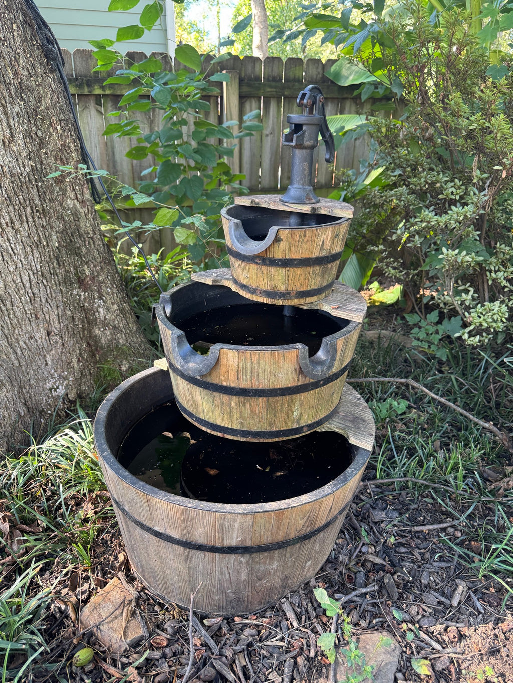Three-tier whiskey barrel fountain with cast-iron-look hand pump set in a garden bed