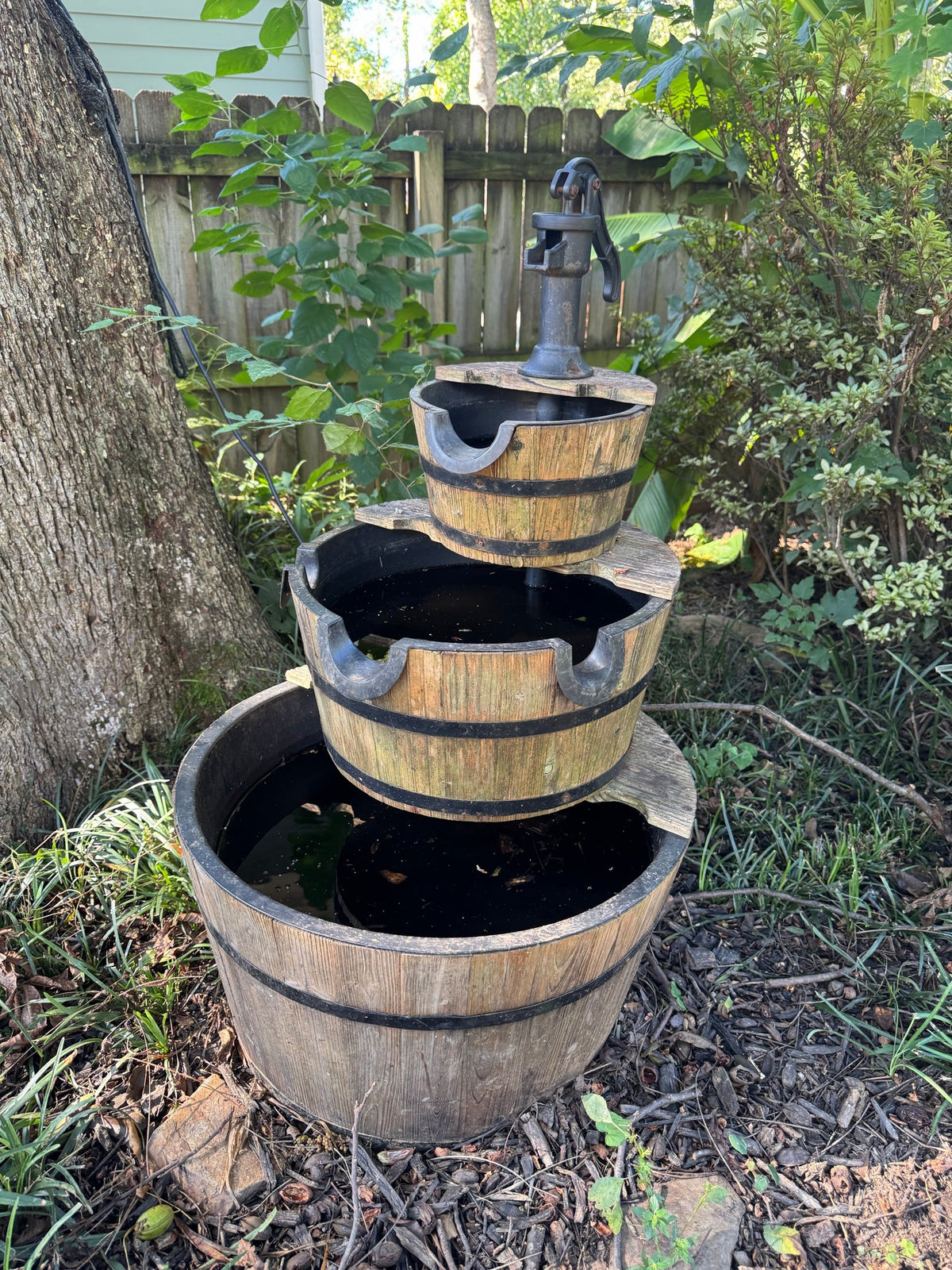 Three-tier whiskey barrel fountain with cast-iron-look hand pump set in a garden bed