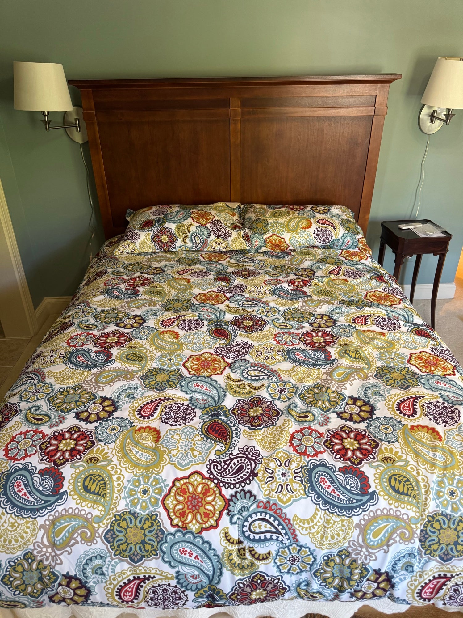 Queen bed with cherry finish panelled headboard, stepped crown molding, colorful paisley patterned bedding and pillows, sage green walls