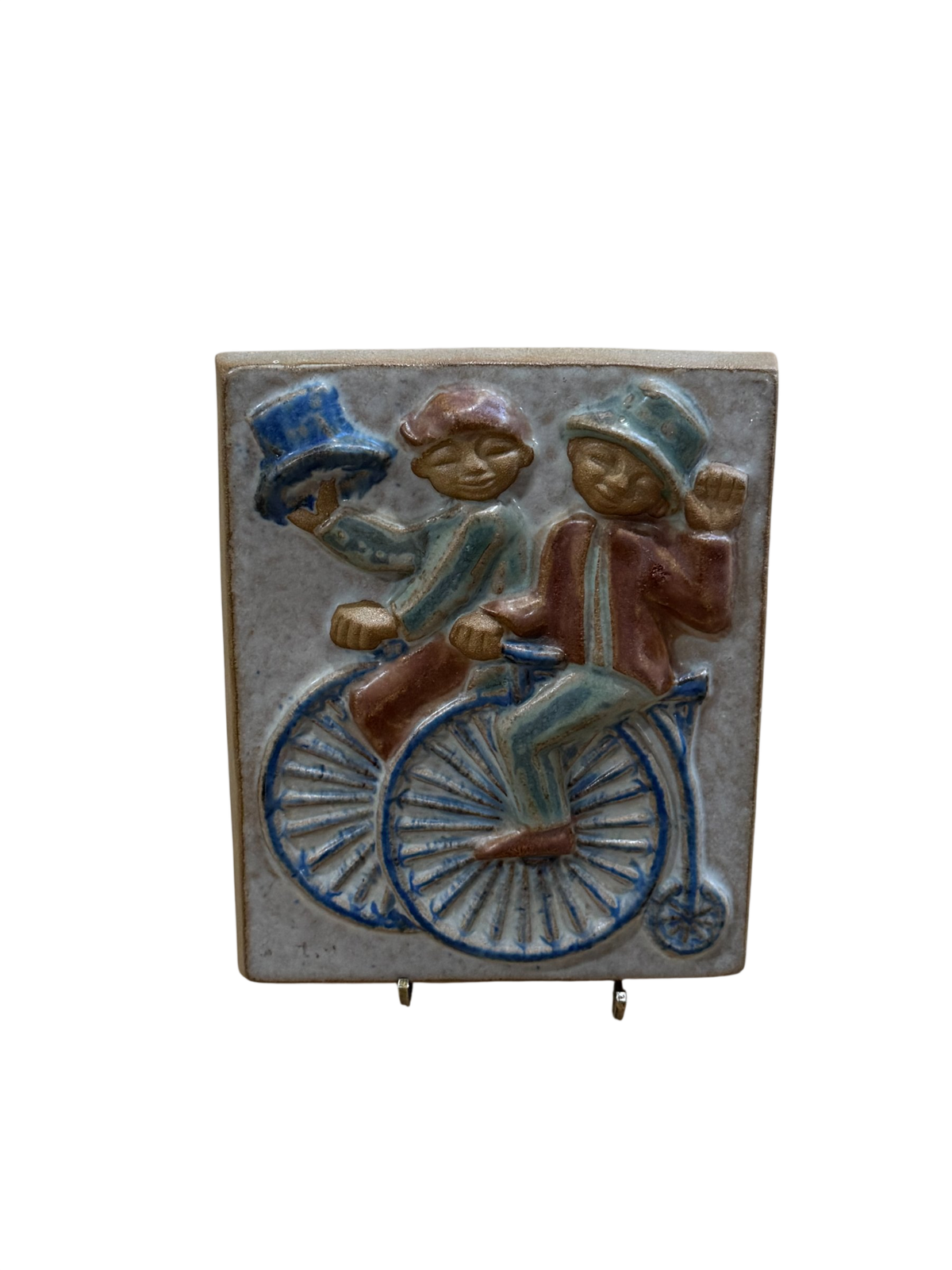 Front view of colored relief tile plaque showing three children on a bicycle with blue wheels, wearing hats and coats in red, blue and green
