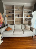 Beige three-seat sofa with rust pillow in front of built-in shelves