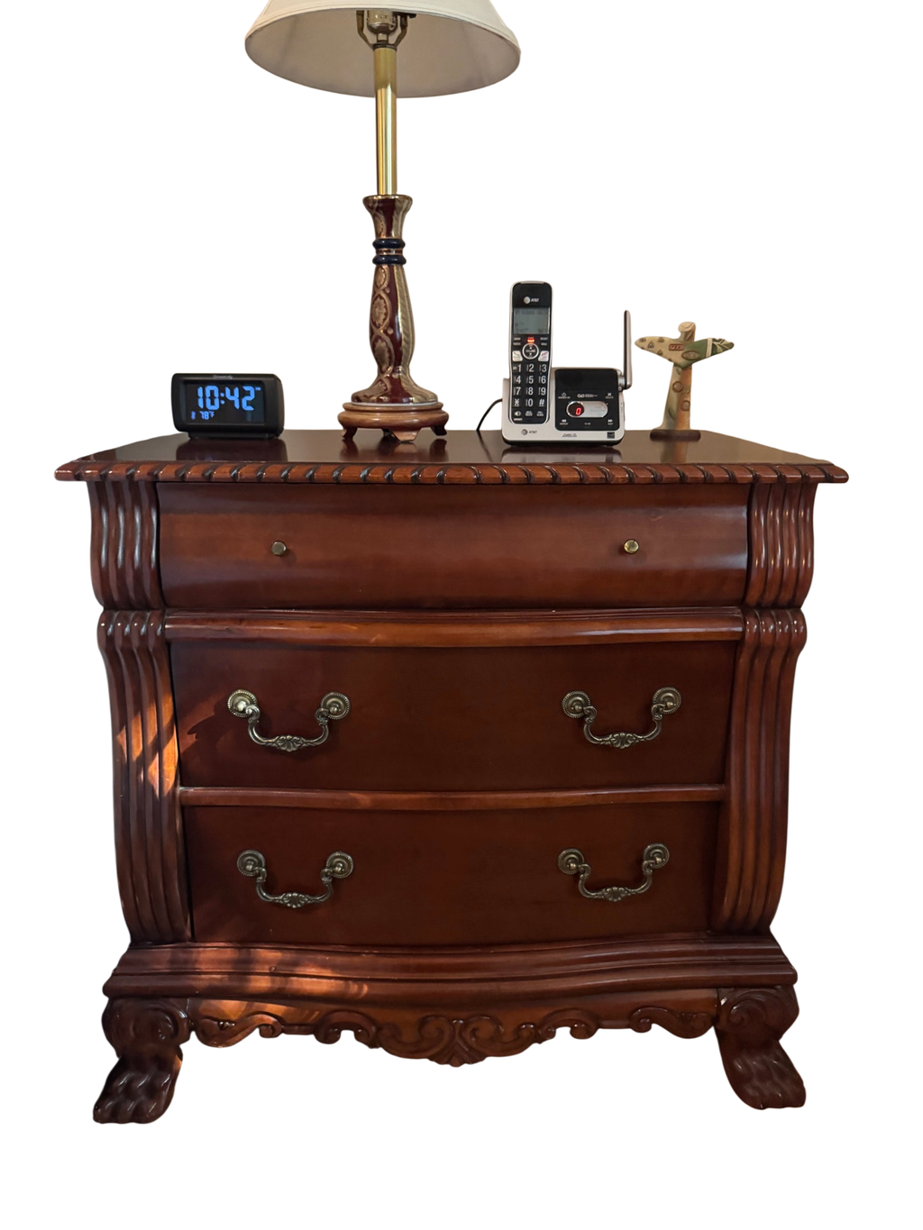 Cherry-finish three-drawer nightstand with carved apron and claw feet, styled with a lamp, clock, cordless phone, and figurine on top