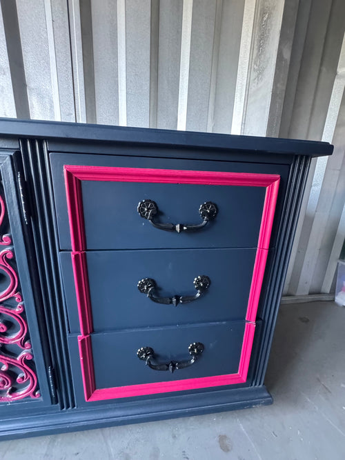 Navy dresser with three drawers, pink frame trim, and black handles, front view on concrete