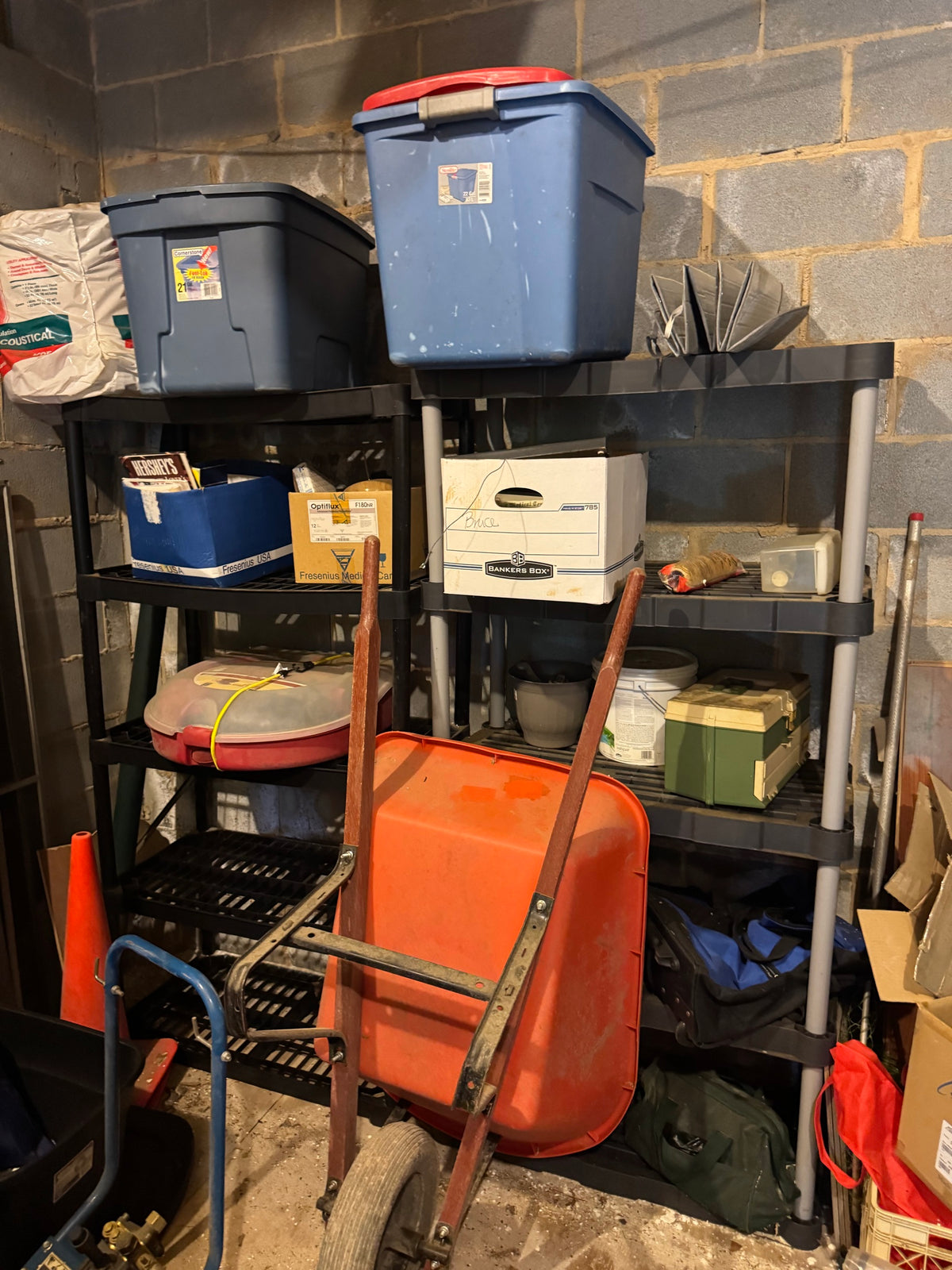 Plastic shelving unit with storage totes, cardboard boxes, a tackle box, wheelbarrow, and an orange traffic cone in a garage
