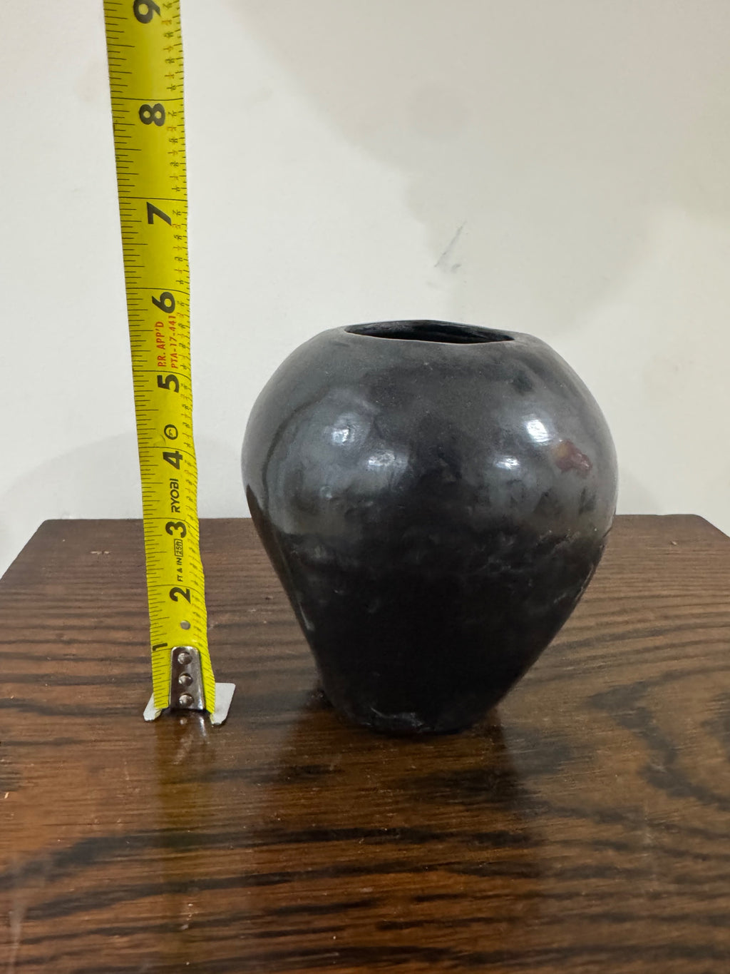 Black pottery vase next to yellow measuring tape showing height of approximately 4-5 inches, positioned on wooden table