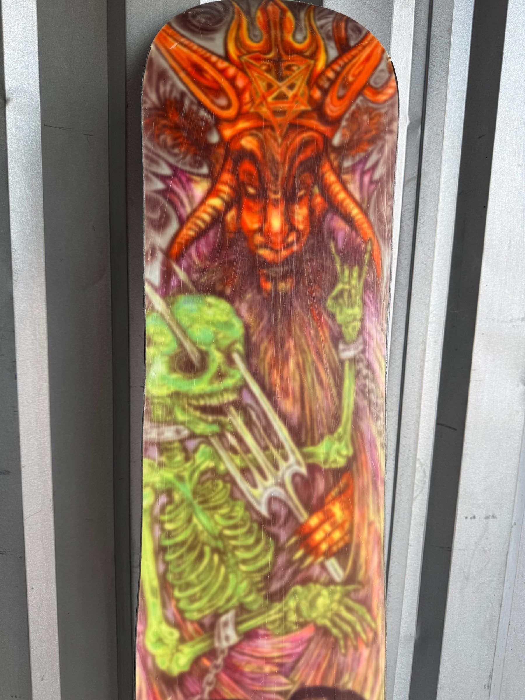 Close-up of snowboard top section featuring vibrant lime green skeleton ribcage and orange demon head with flame details