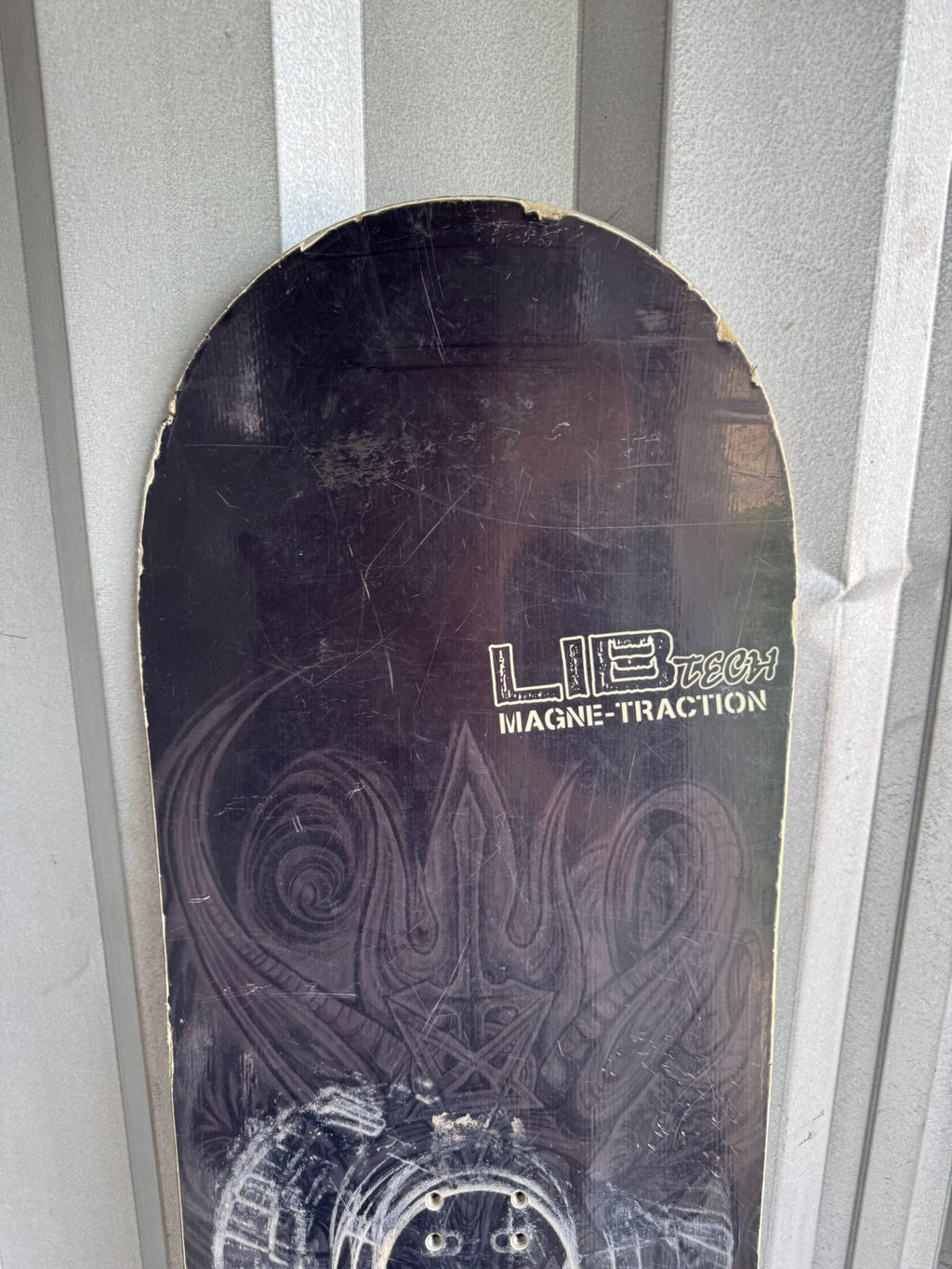 Snowboard base showing Lib Tech Magne-Traction branding, circular binding mounting holes and black surface with embossed pattern details