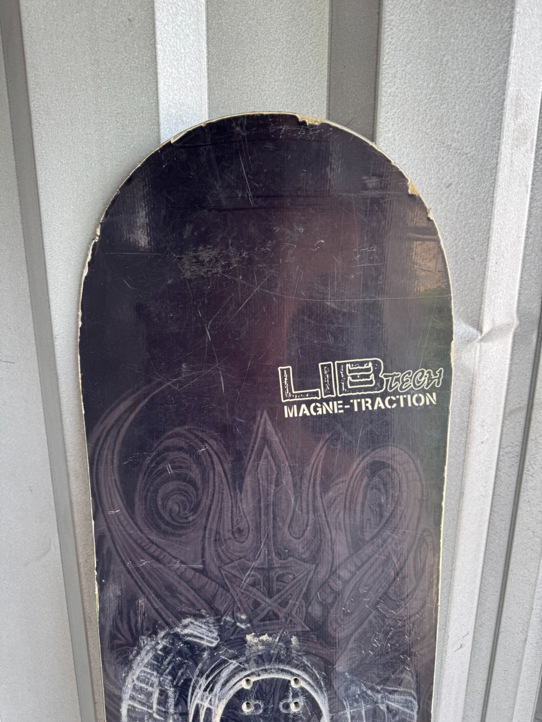 Snowboard base showing Lib Tech Magne-Traction branding, circular binding mounting holes and black surface with embossed pattern details