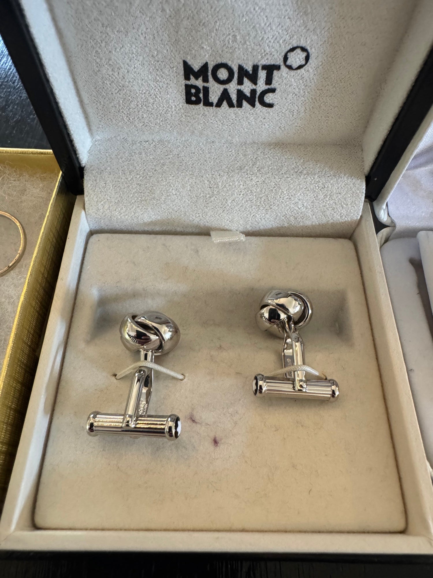 Montblanc branded presentation box open, displaying pair of silver-toned knot cufflinks on cream velvet interior