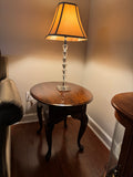 Round cherry finish side table with a single drawer and cabriole legs in a living room corner, topped with a crystal-style table lamp