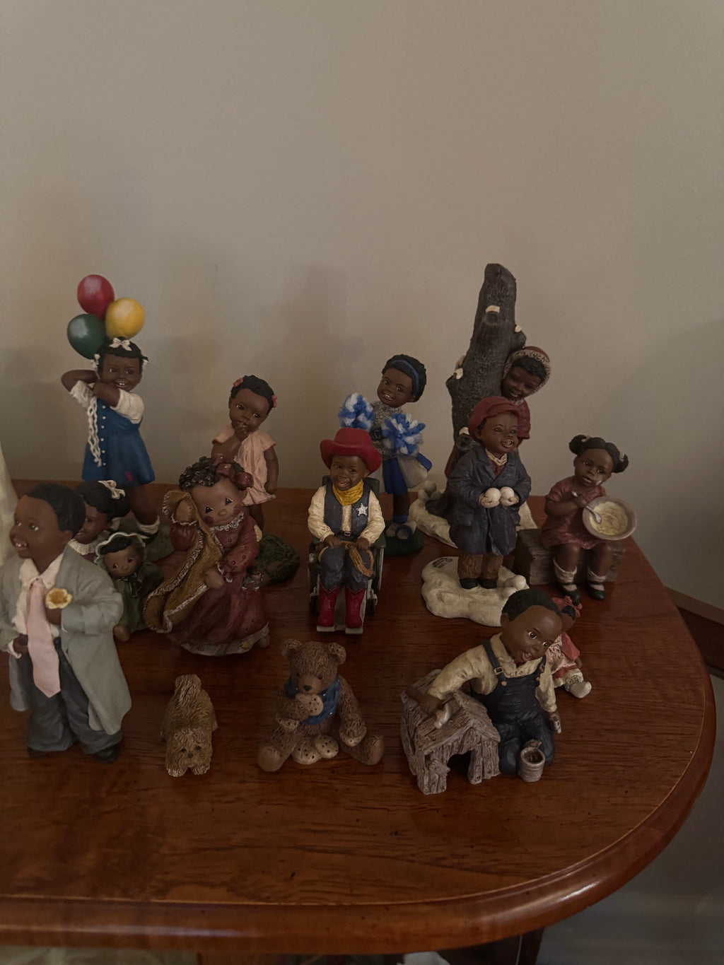 Grouping of All God's Children figurines on a wood table including a girl with balloons, a cowboy in a wheelchair, and teddy bear figures
