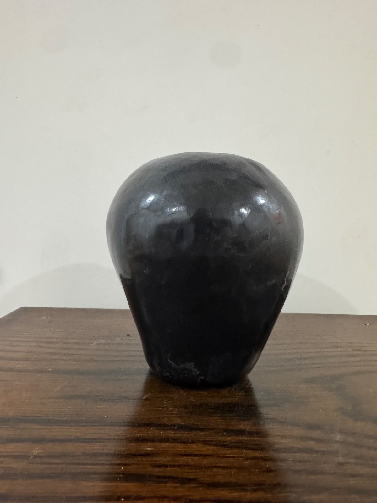 Black pottery vase shown from side angle on wooden surface with white wall background, demonstrating spherical shape and glossy finish