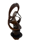 Abstract wooden sculpture displayed on black hexagonal pedestal, carved organic forms with circular voids, dark wood finish, studio lighting