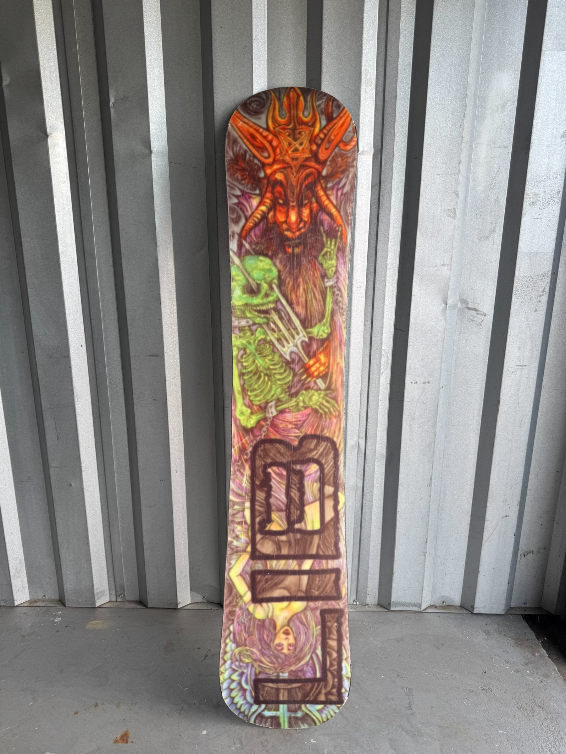 Snowboard standing upright against corrugated metal background, colorful skeleton and demon artwork visible on top half