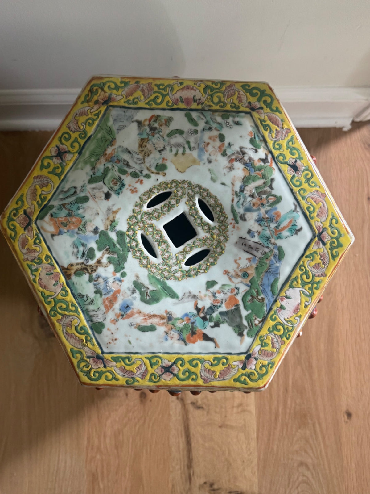 Top-down view of hexagonal stool showing yellow scrollwork border, painted figures and landscape scene, ventilation holes