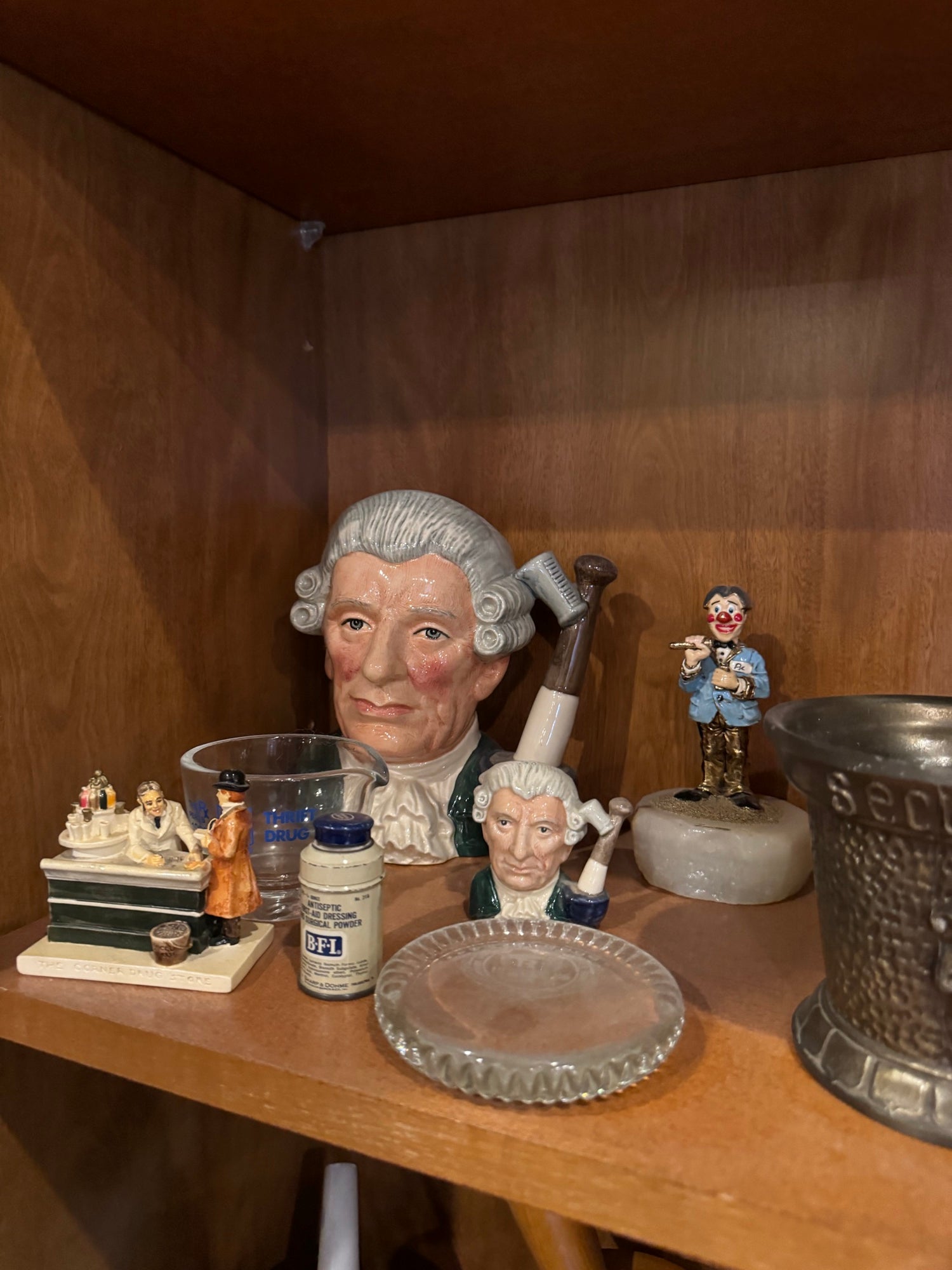 Character jug displayed on wooden shelf alongside various collectibles including smaller jugs, decorative plate, and metal vessel