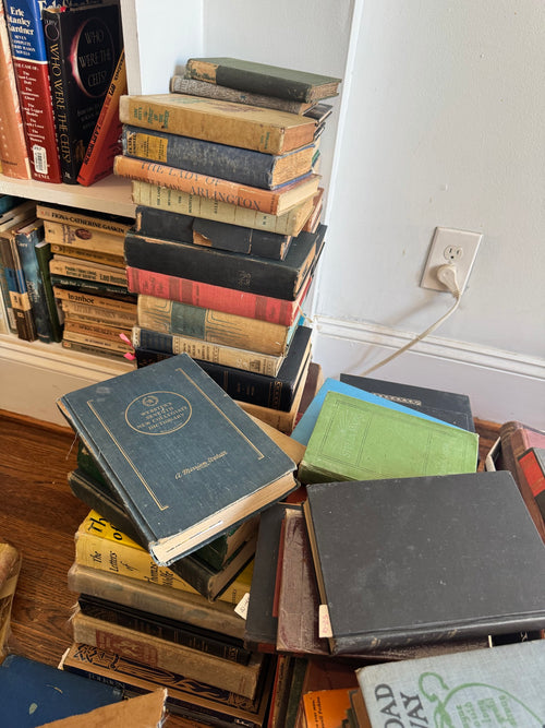 Tall stack of vintage hardcover books arranged vertically on wooden floor, spine titles visible, ranging from tan to dark covers