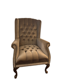 Champagne satin tufted wingback chair with nailhead trim and curved wood legs in a corner near a window.