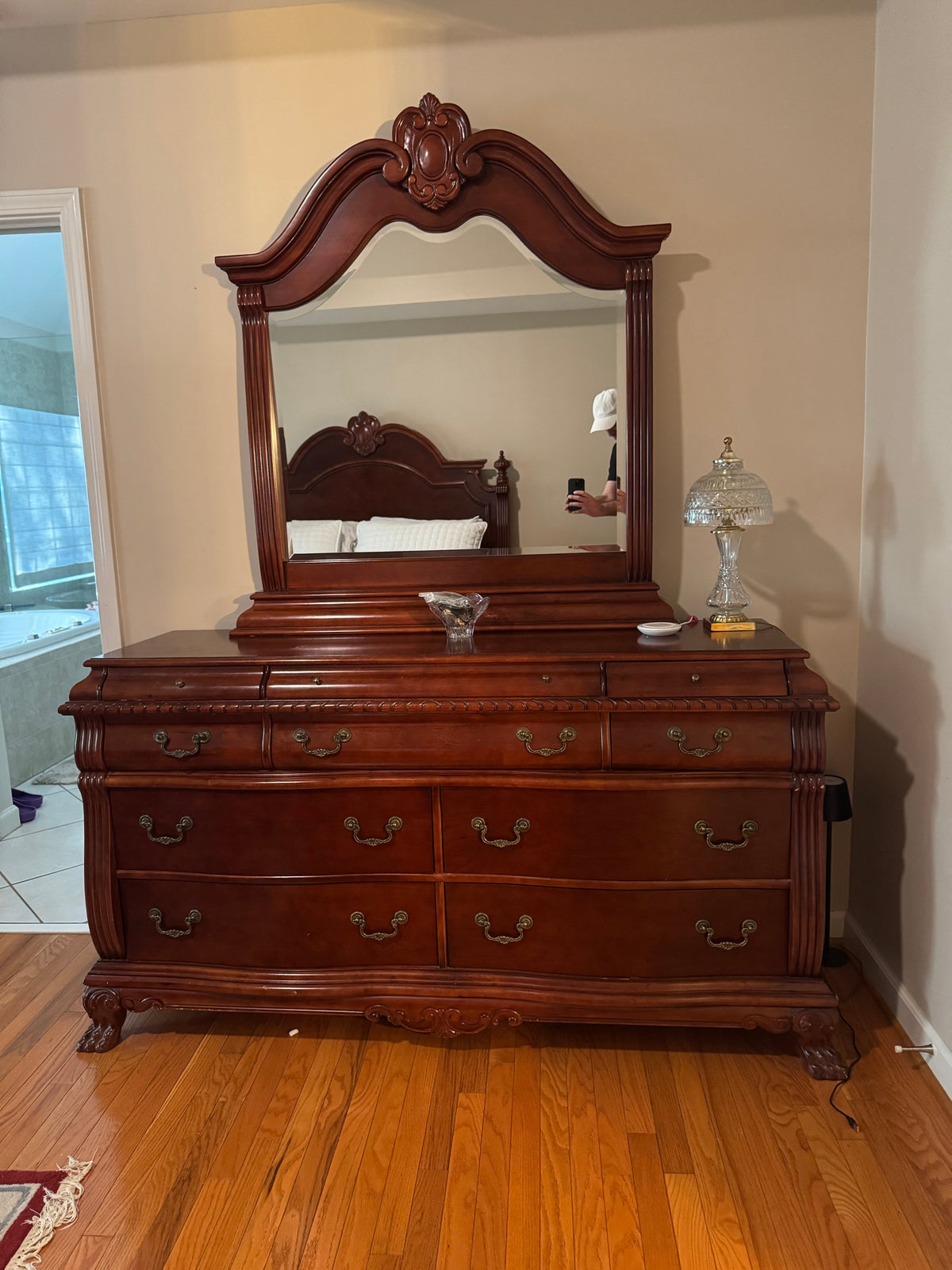 Cherry-finish wood triple dresser with attached arched mirror, brass drawer pulls, and carved details in a bedroom
