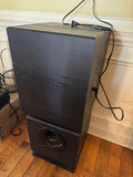 Black subwoofer with textured finish, standing upright on wooden floor against cream wall